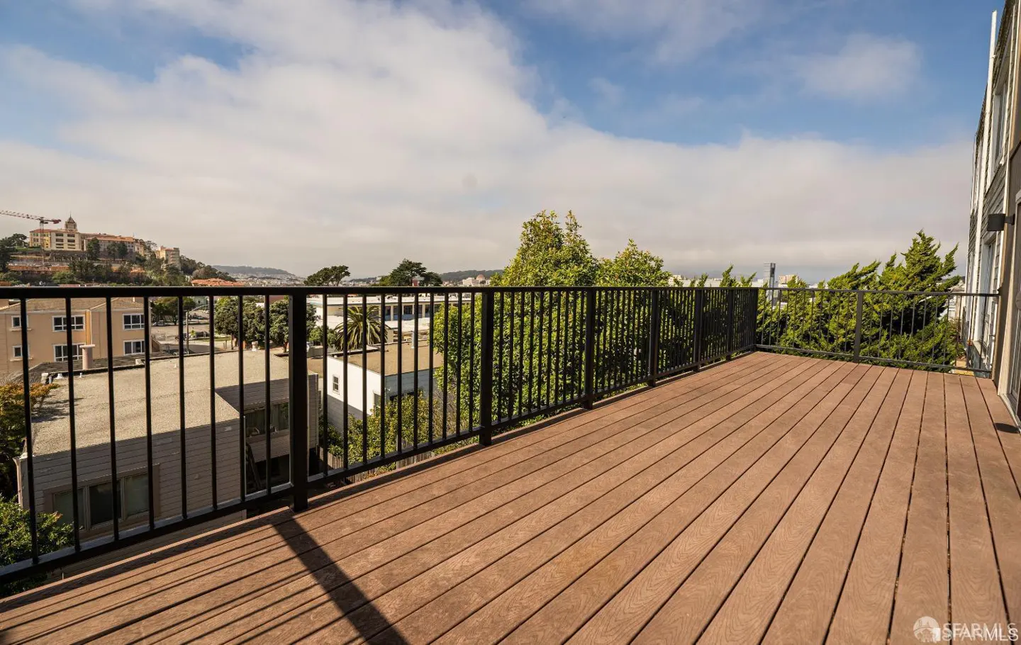 A wood deck with black railings overlooks a city with trees and buildings under a blue sky.