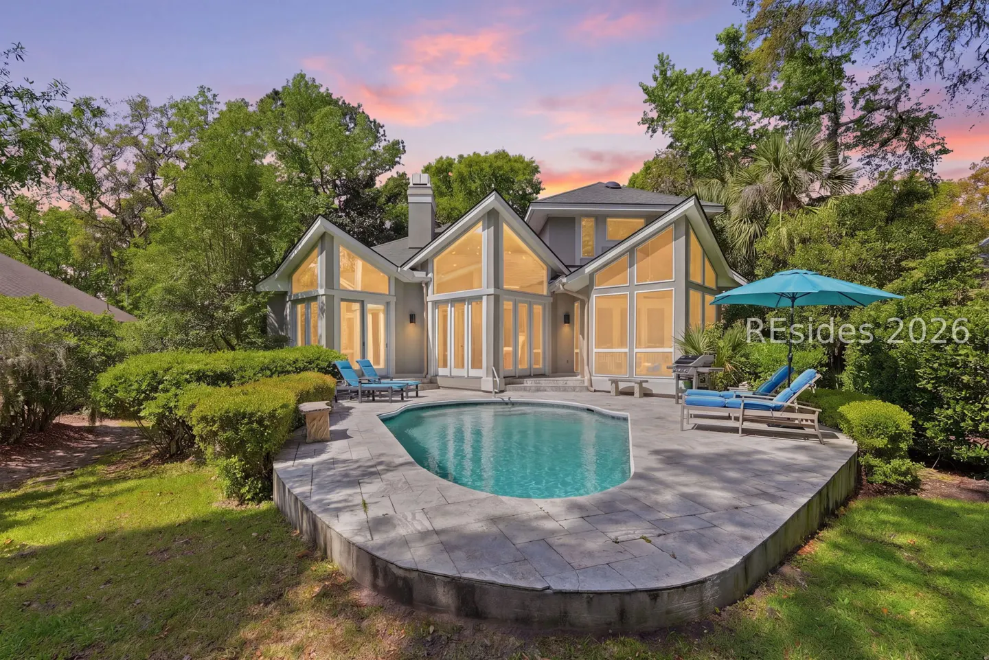 Backyard view of a gray house with a pool, patio, lounge chairs, and large windows. Trees and a colorful sunset are in the background.