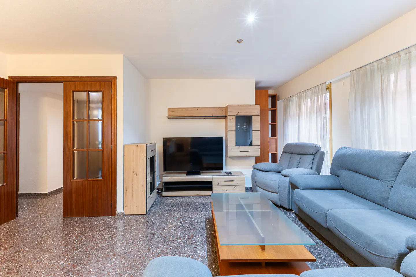 Living room with a blue sofa, glass coffee table, TV, and wood furniture. A doorway is visible on the left.
