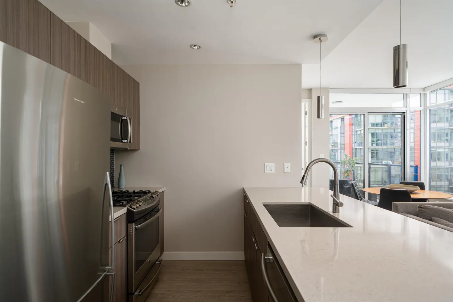 Modern kitchen with stainless steel appliances, wood cabinets, and a white countertop island with a sink. Large windows offer a city view.