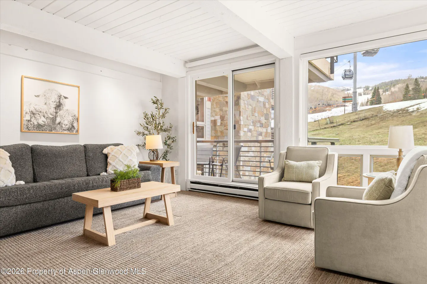 Living room with gray sofa, two beige chairs, and a light wood coffee table. A ski lift is visible through the window.