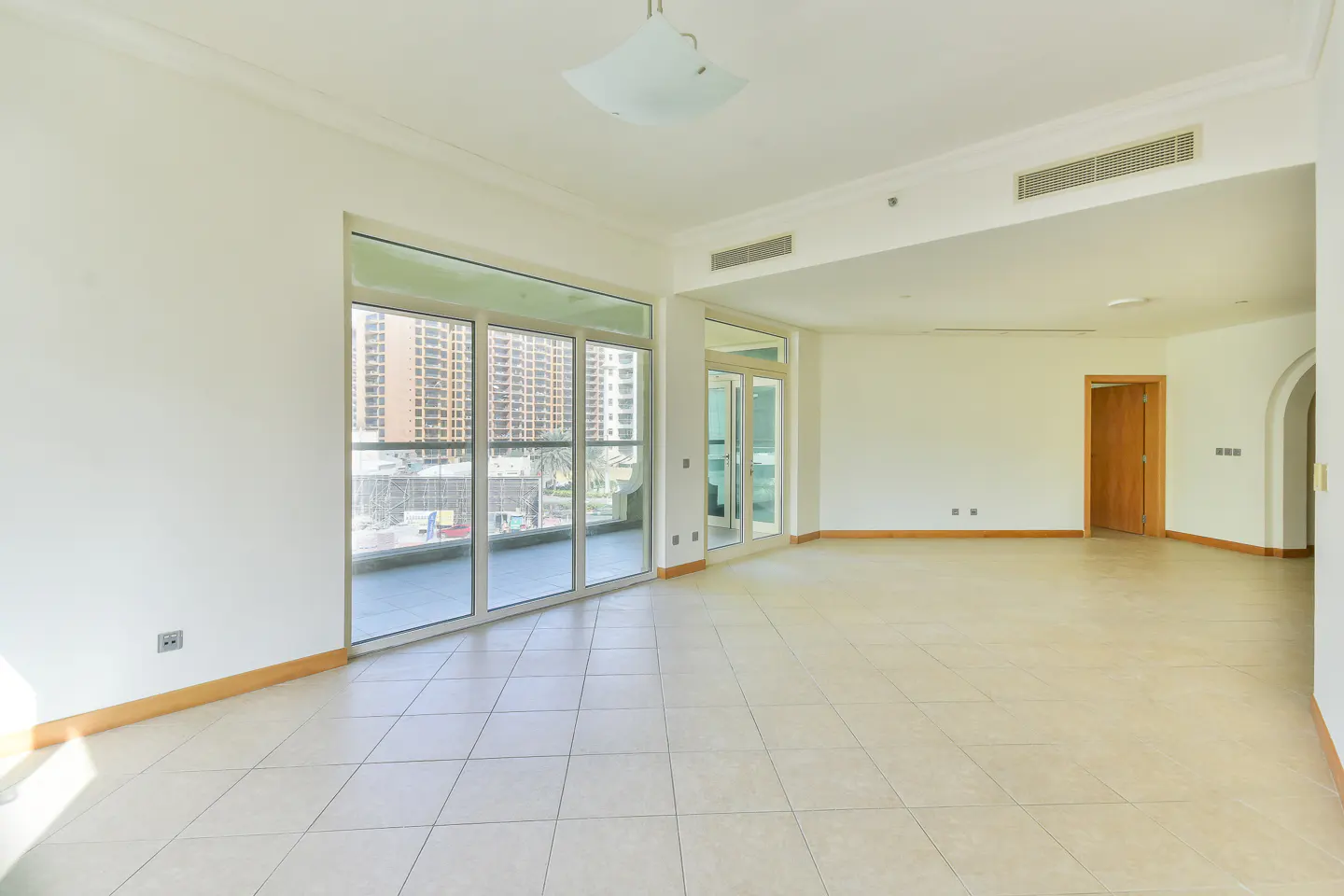 Bright, empty room with white walls, tile floor, and large glass doors to a balcony with a city view.
