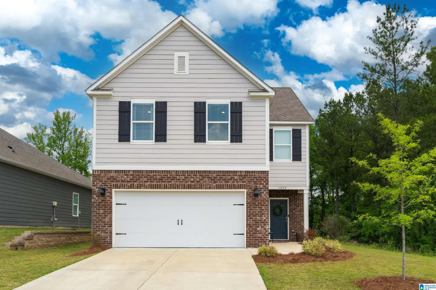 Two-story suburban home with light gray siding, brick lower level, white garage door, and dark shutters under a blue sky.