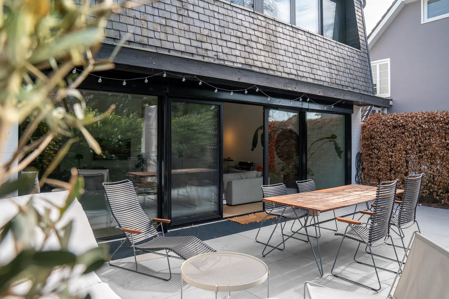 Outdoor patio with a wood table, chairs, and lounge seating. Sliding glass doors lead to the home's interior. String lights hang above.
