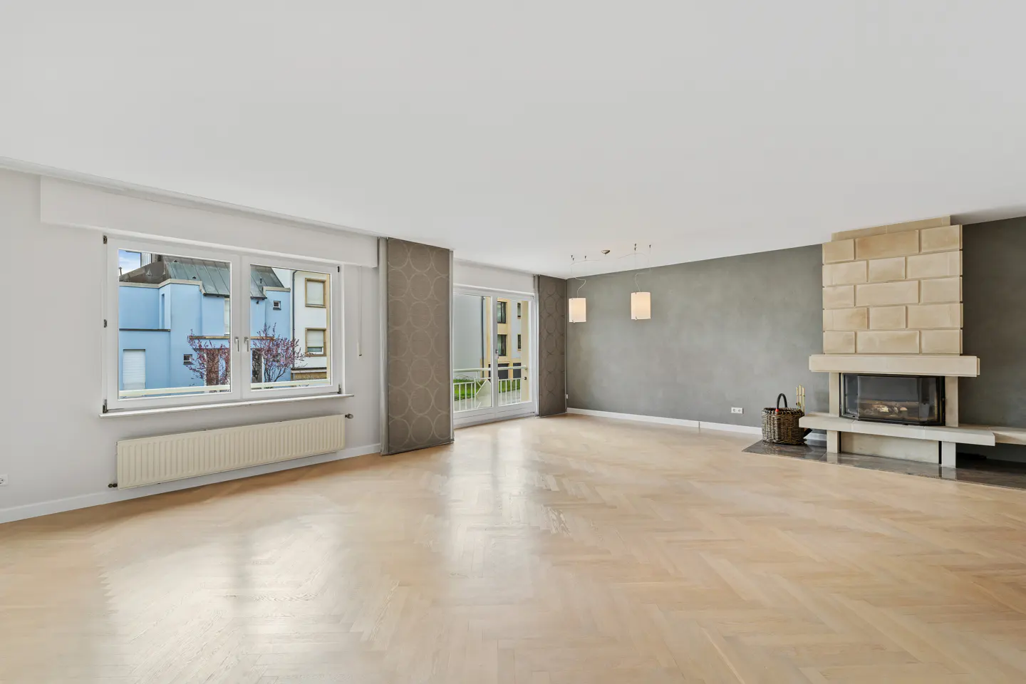 Bright, empty living room with herringbone wood floors, stone fireplace, and large windows. Gray accent wall.