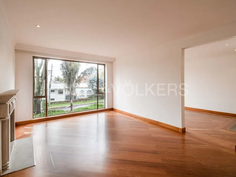 Bright, empty living room with hardwood floors, white walls, and a large window overlooking a tree-lined street. A stone fireplace is on the left.