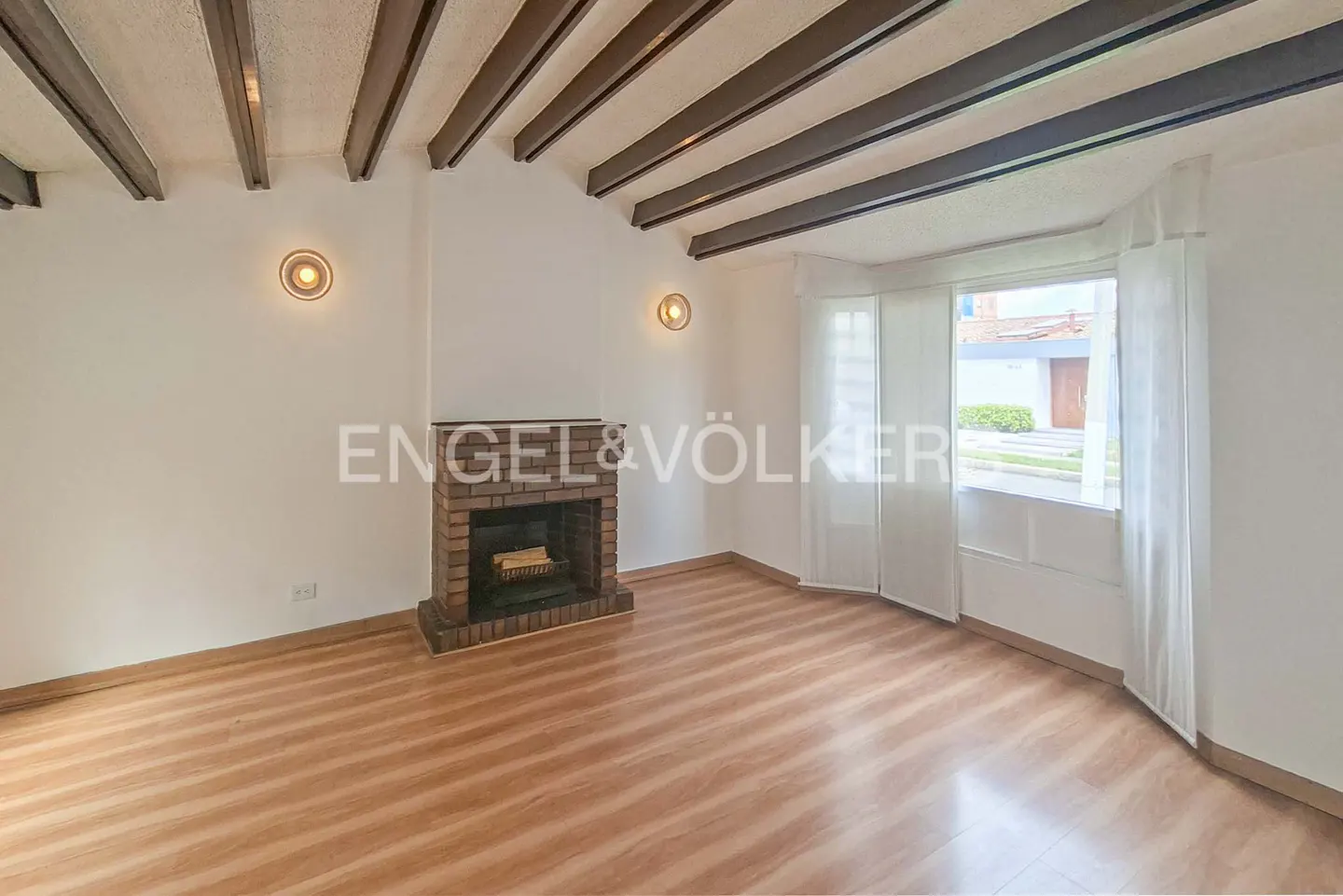 Living room with wood-look floors, exposed ceiling beams, a brick fireplace, and large windows with white curtains.