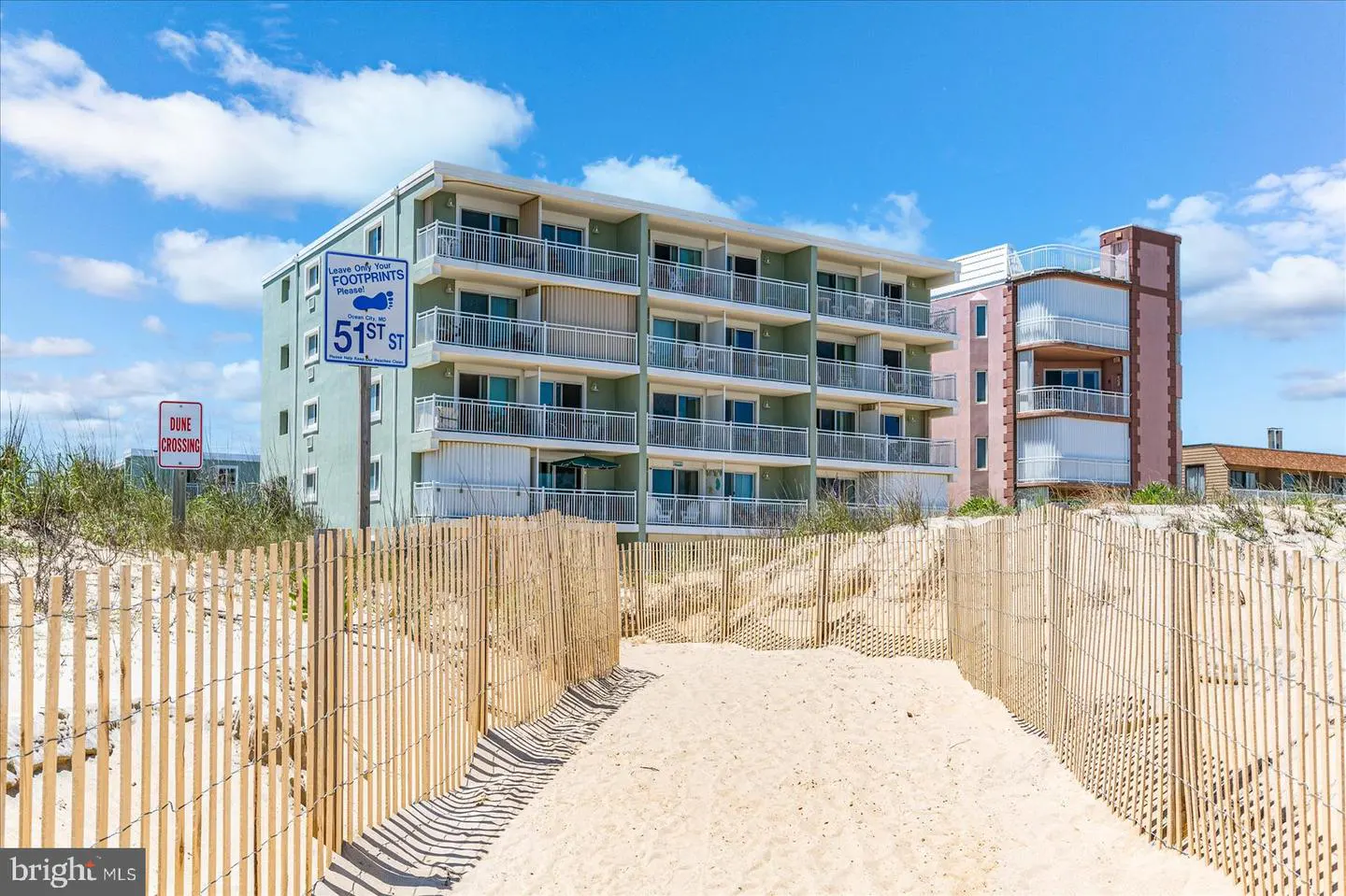 Beachfront condo with a sandy path leading to the building. The building is light green with white balconies. Blue sky.