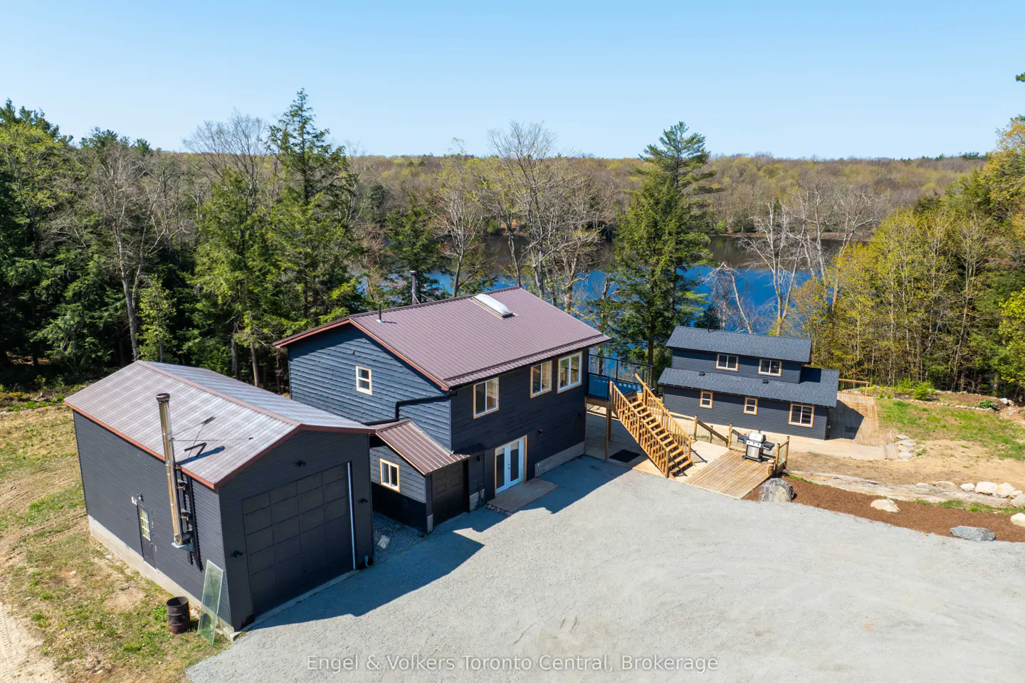 Aerial view of a dark blue house with a brown roof, a garage, and a guest house near a lake surrounded by trees.