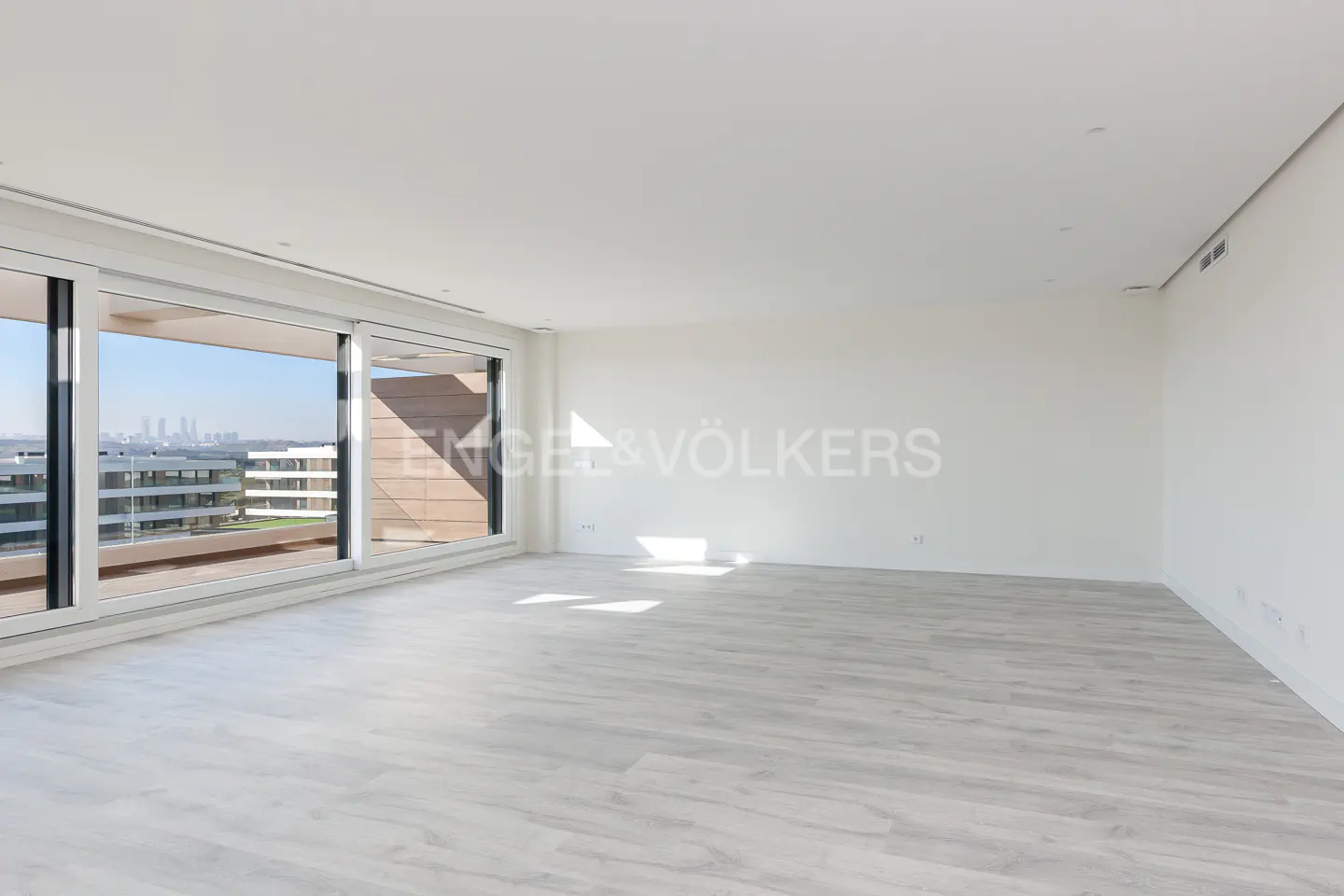 Bright, empty room with white walls, light wood floors, and a large window with a city view.