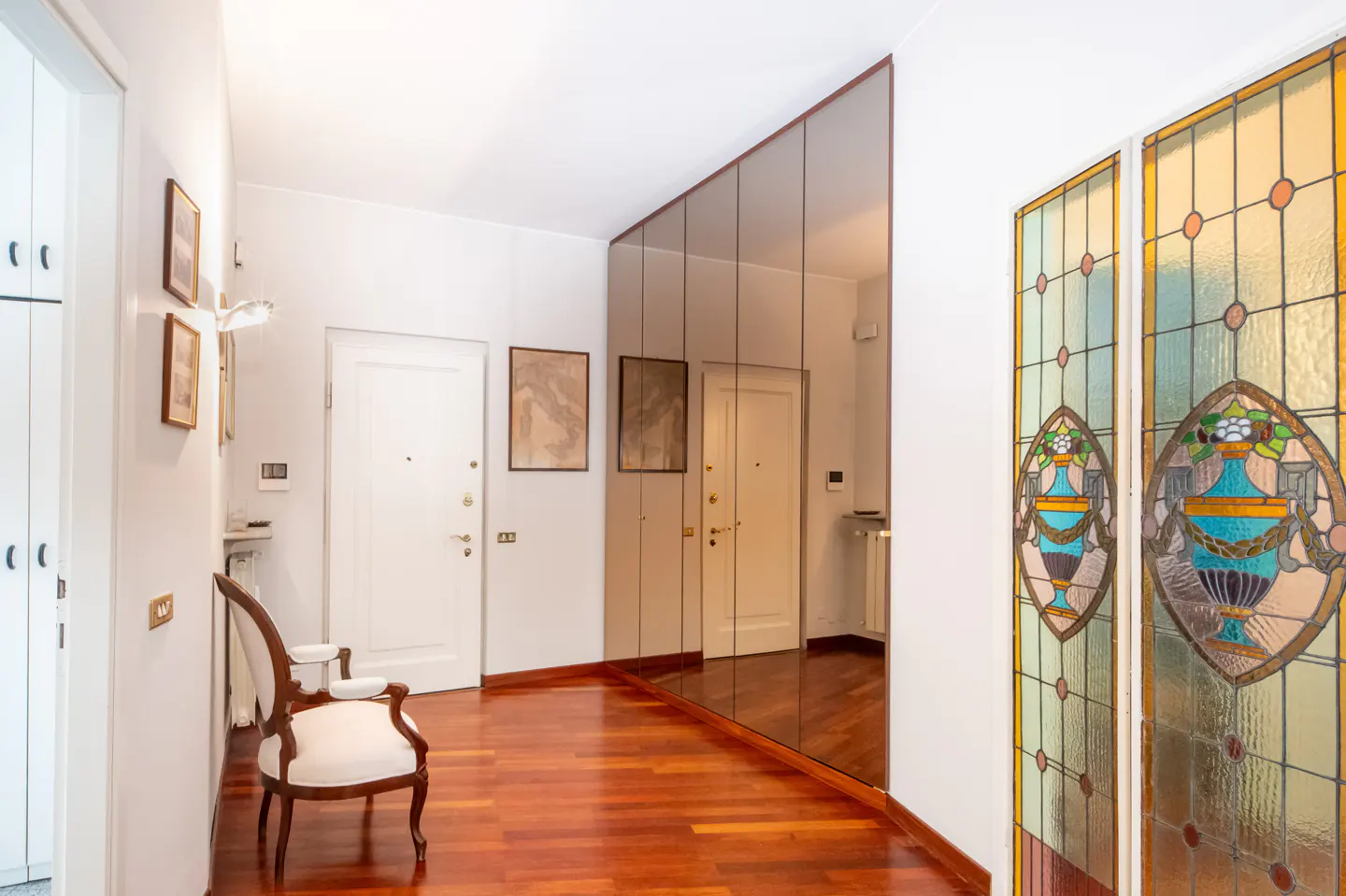 Hallway with hardwood floors, white walls, and a mirrored wall. Stained glass panels and a white chair add detail.