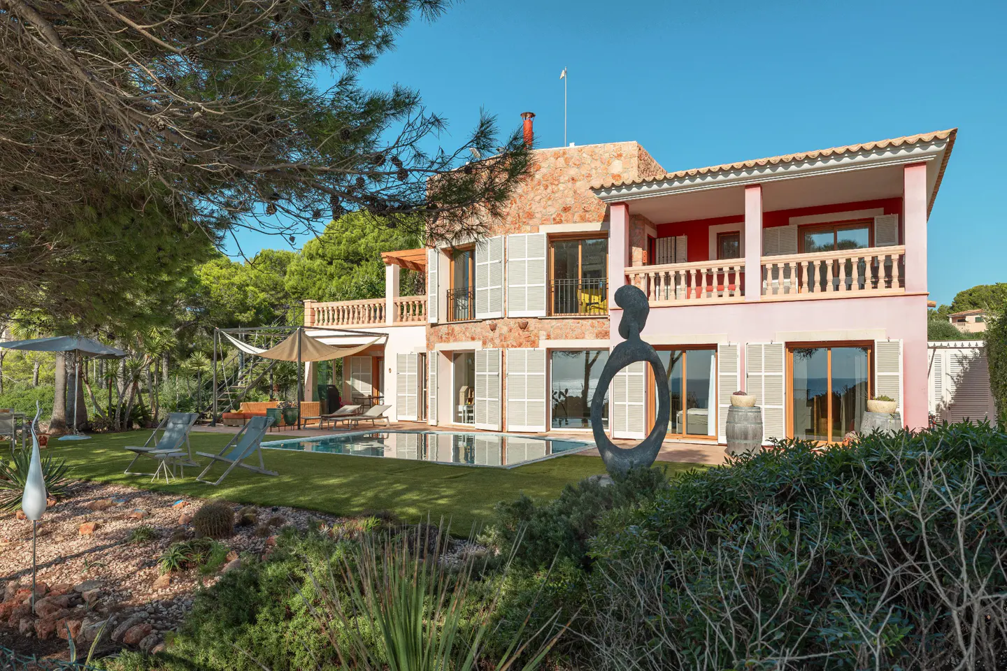 Two-story pink house with stone accents, white shutters, and a balcony overlooking a pool and green lawn. A modern sculpture stands in the foreground.