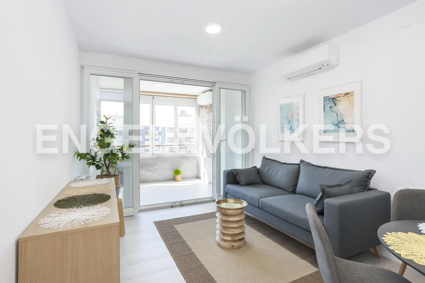 Bright living room with a grey sofa, wooden side table, area rug, and sliding glass doors leading to a balcony.