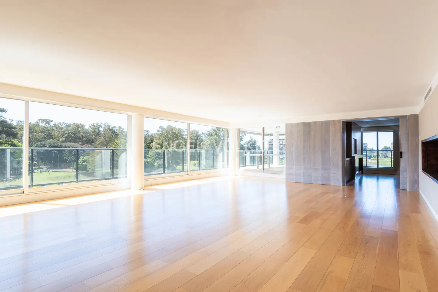 Bright, empty living room with hardwood floors and floor-to-ceiling windows overlooking a green landscape.