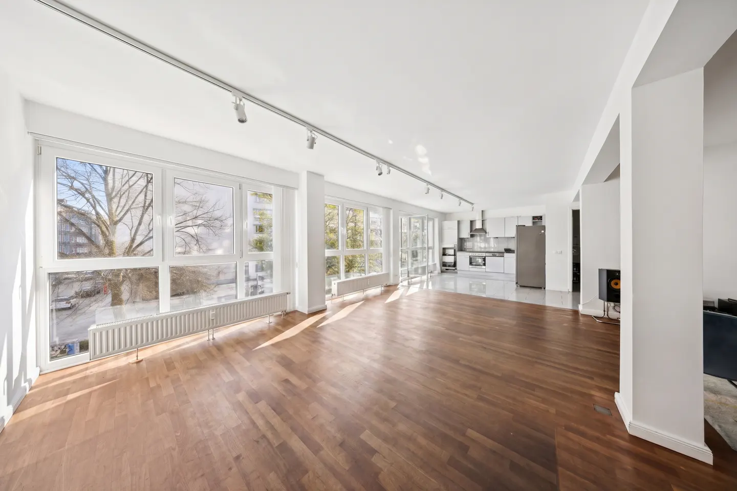 Bright, open-concept apartment with hardwood floors, white walls, and track lighting. Large windows offer city views. Kitchen in background.