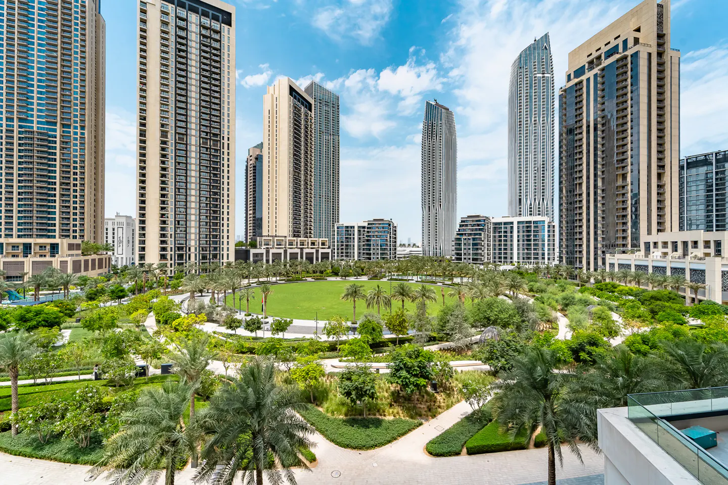 Cityscape view of modern high-rise buildings surrounding a green park with palm trees under a blue sky.