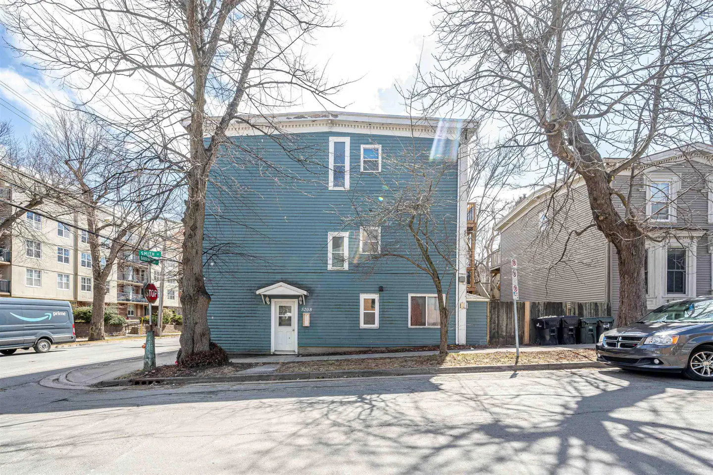 Exterior view of a three-story blue house with white trim, framed by bare trees on a sunny day.