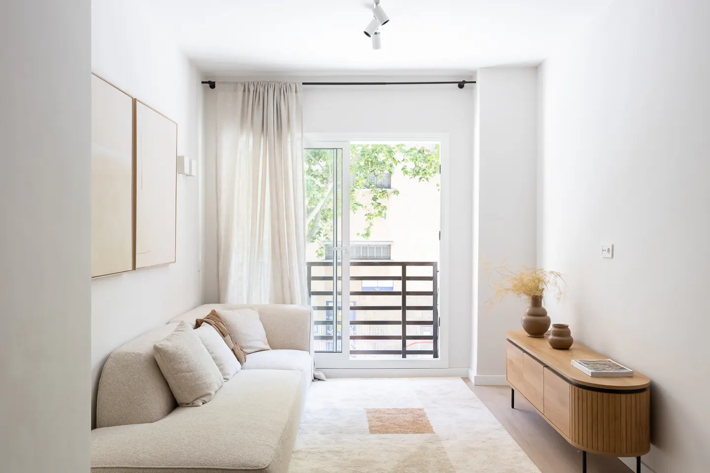 Bright, minimalist living room with a beige sofa, wooden sideboard, and a glass door leading to a balcony with trees.