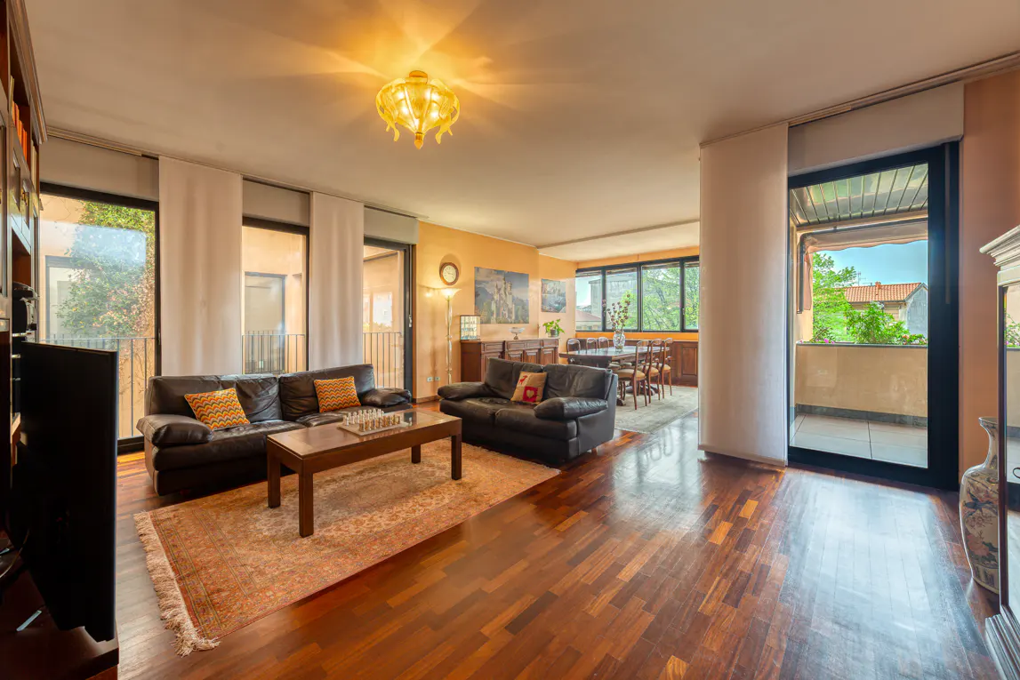 A warm-toned living room with dark wood floors, two black leather sofas, and a dining area visible in the background. Balcony access is on the right.