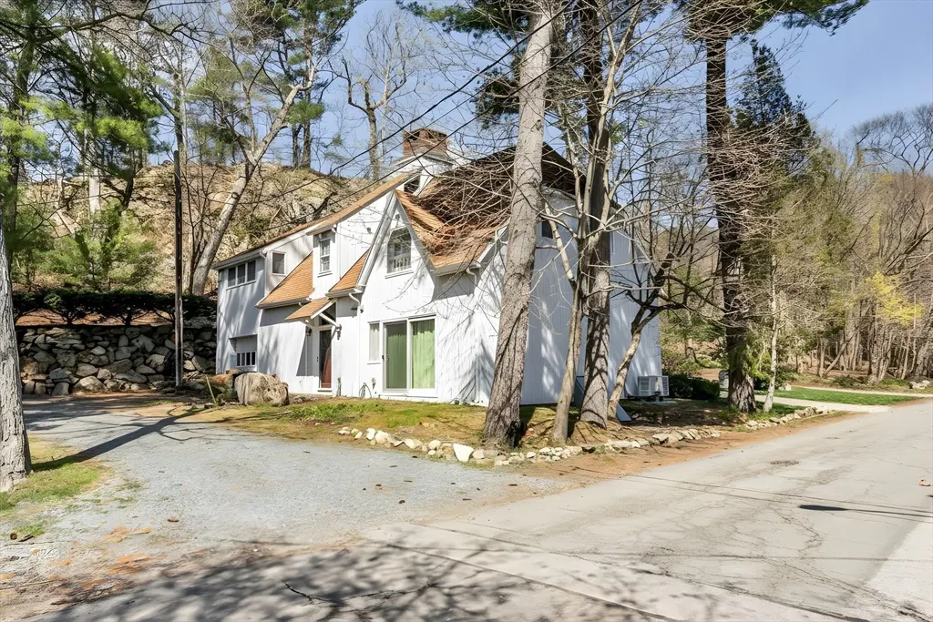 Exterior view of a white house with a brown roof, surrounded by trees and a stone wall. A gravel driveway leads to the house.