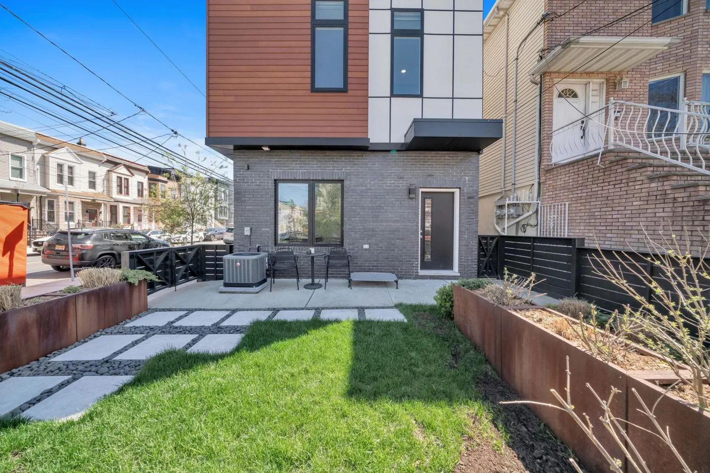 Backyard view of a modern townhouse with a small lawn, patio, and metal planters on a sunny day.