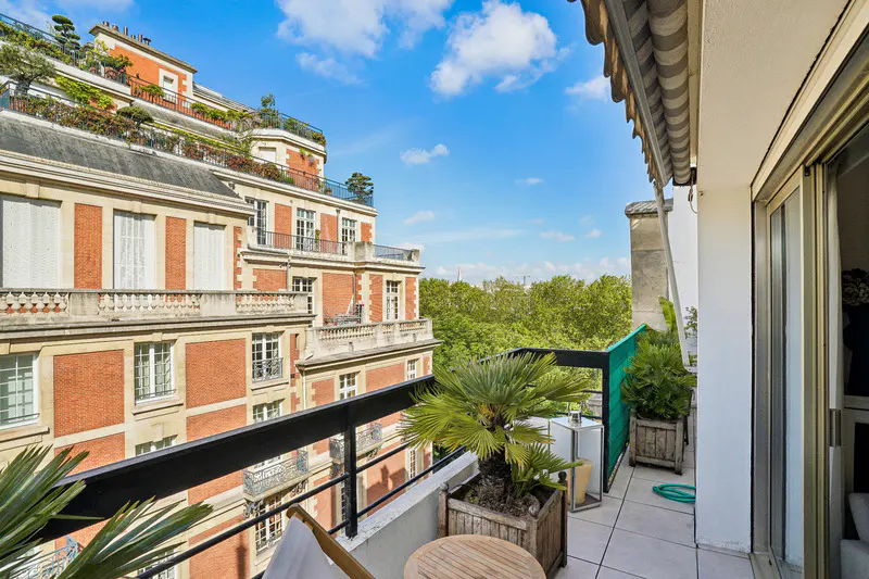 Balcony view of a Parisian apartment with potted plants, a table, and a chair. A red brick building and green trees are visible in the background.