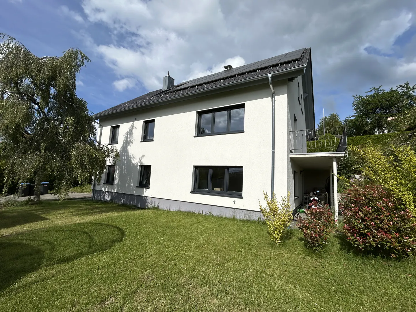 Two-story white house with a dark roof, solar panels, and a grassy yard under a cloudy blue sky.