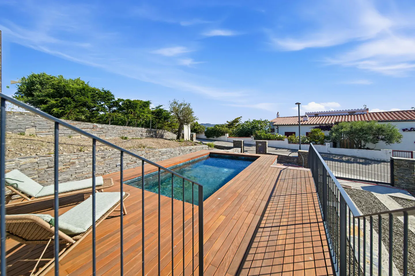 Outdoor pool with wooden deck, lounge chairs, and metal railing. Houses and blue sky in the background.