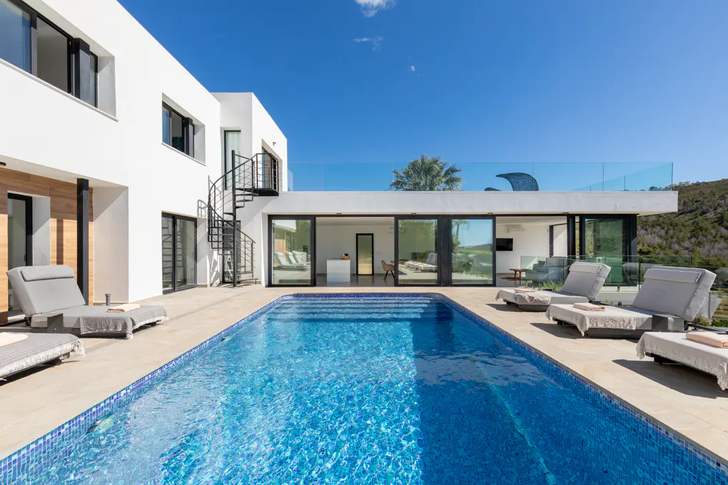 Modern white house with a blue tiled pool, lounge chairs, and a black spiral staircase under a clear blue sky.