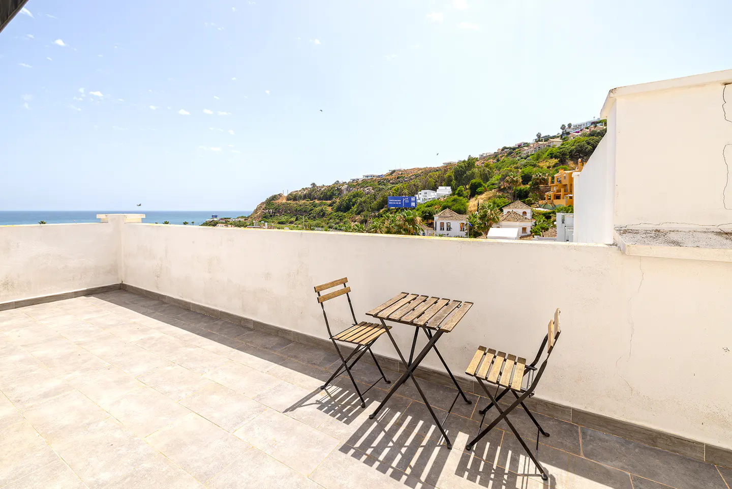 Balcony view with a small wooden table and two chairs, overlooking the ocean and a green hillside under a bright sky.