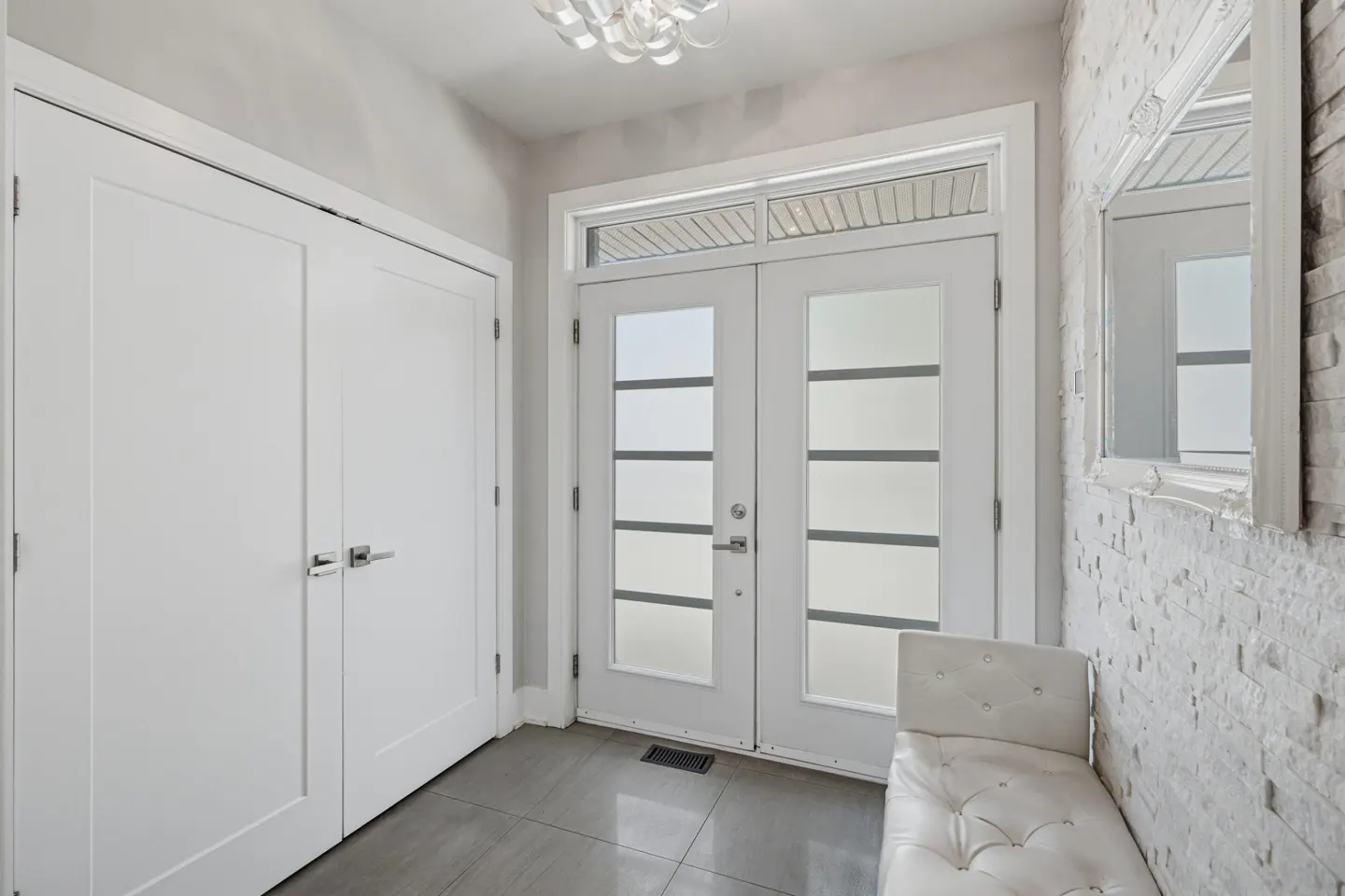 Bright entryway with white double doors, a tufted bench, a stone accent wall, and a large mirror on grey tile flooring.