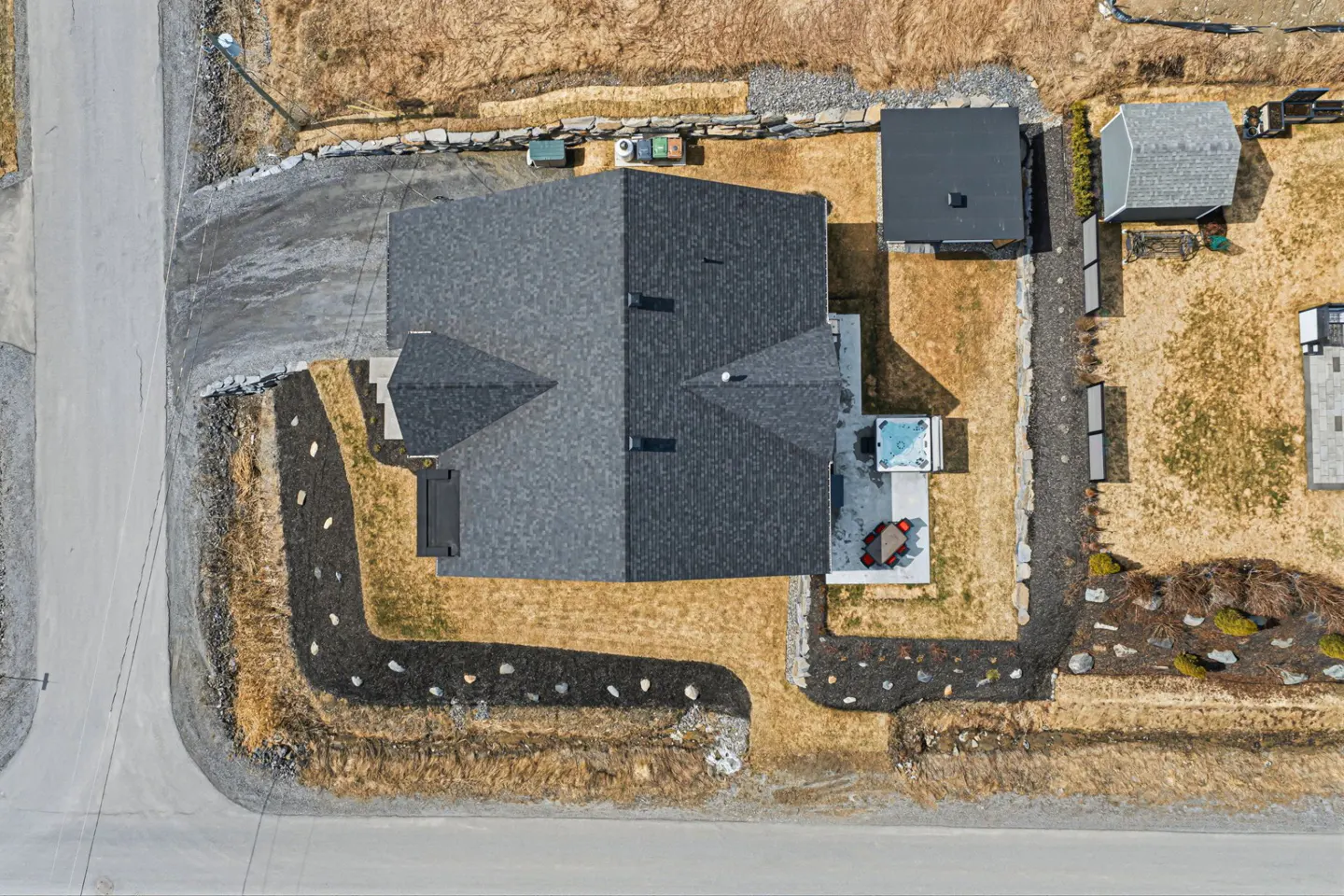Aerial view of a gray roofed house with a hot tub, patio furniture, and a shed in a brown grassy yard.