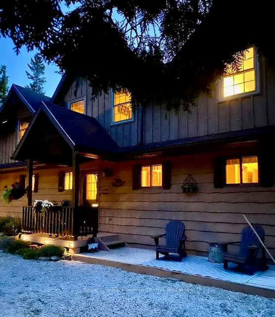 Exterior view of a two-story house with a porch and Adirondack chairs at dusk. Windows glow with warm light.