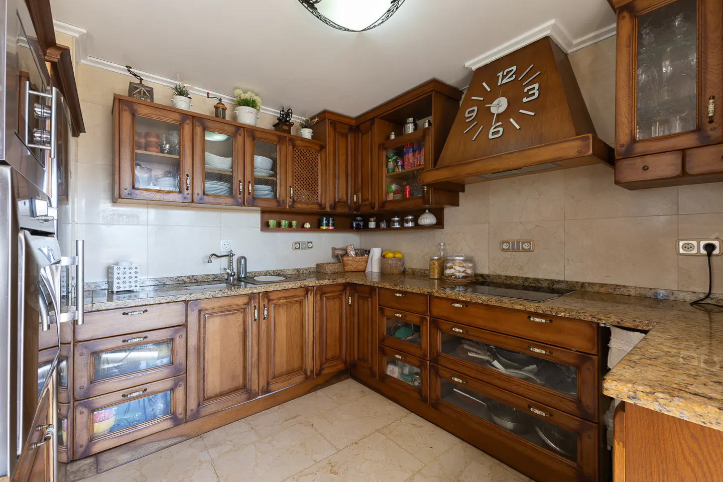 A kitchen with wooden cabinets, granite countertops, and a large clock on the range hood.