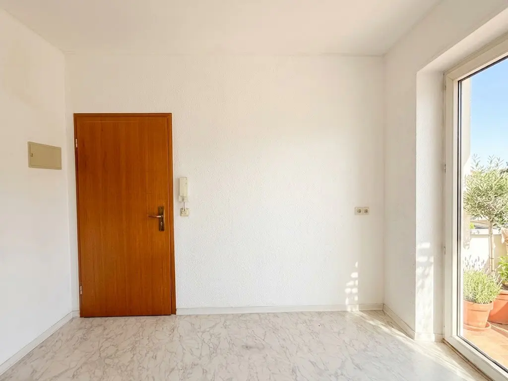 Bright, empty room with white walls, marble floor, and a wooden door. A glass door leads to a balcony with potted plants.