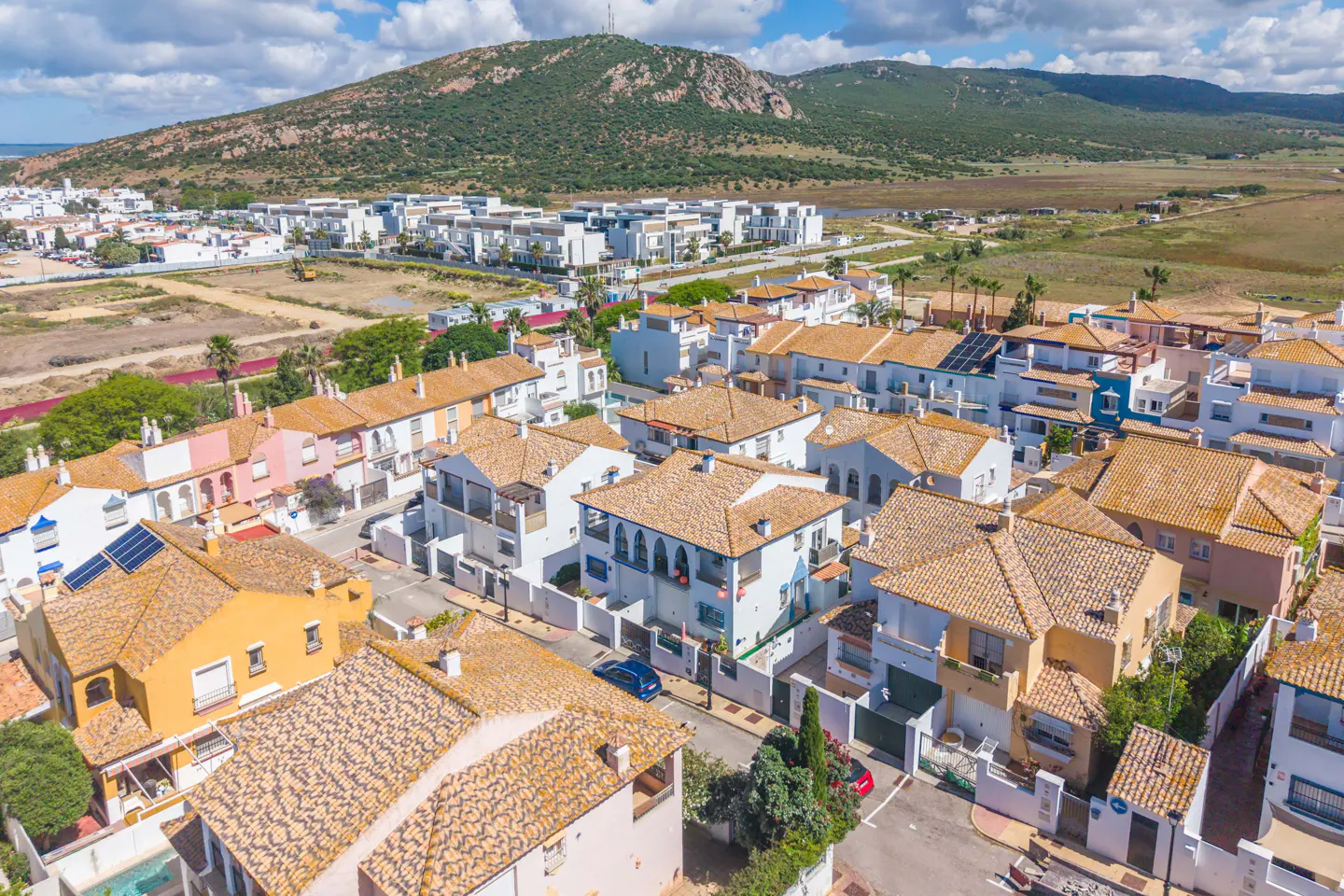 Aerial view of a residential area with terracotta-roofed houses, white walls, and a mountain backdrop under a partly cloudy sky.