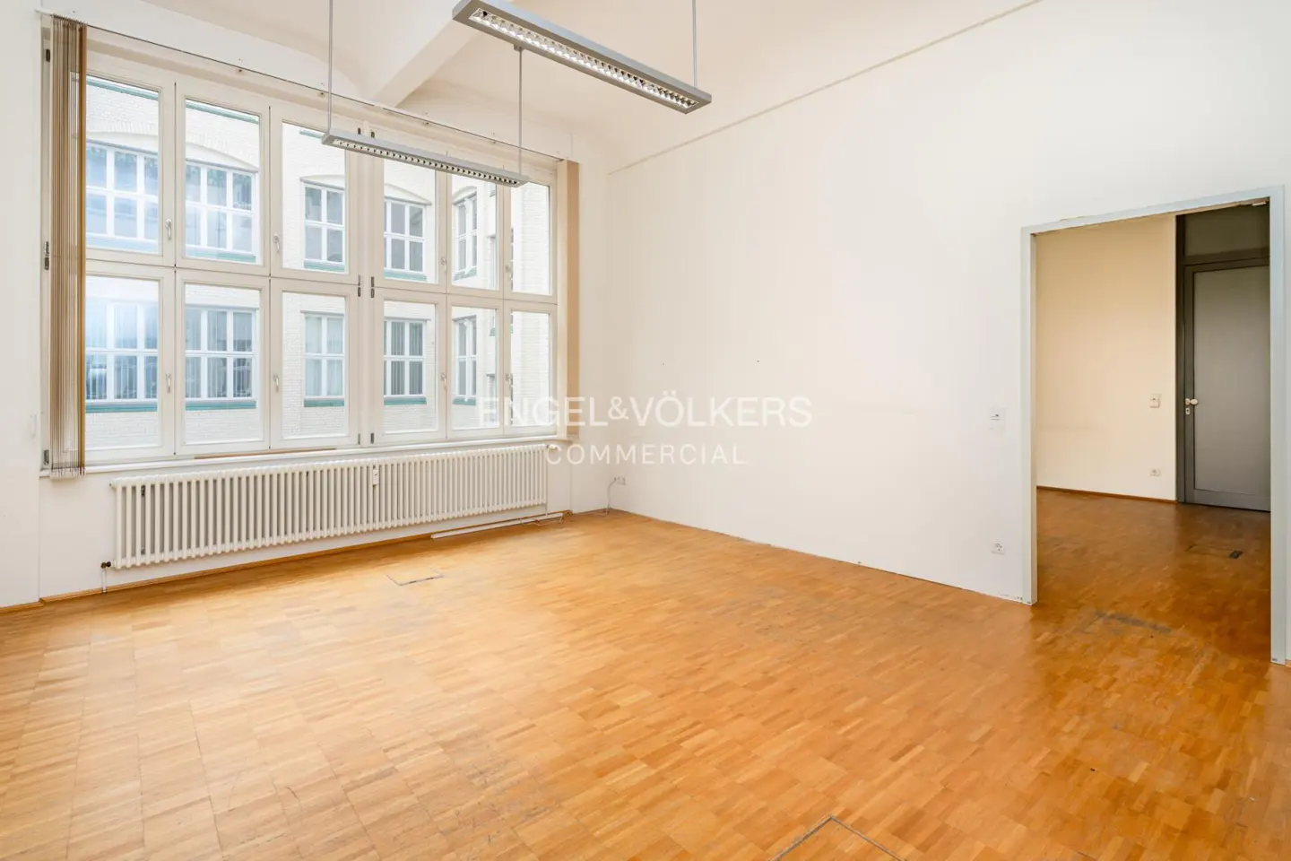 Empty office with light wood parquet flooring, white walls, large windows, and a doorway leading to another room.