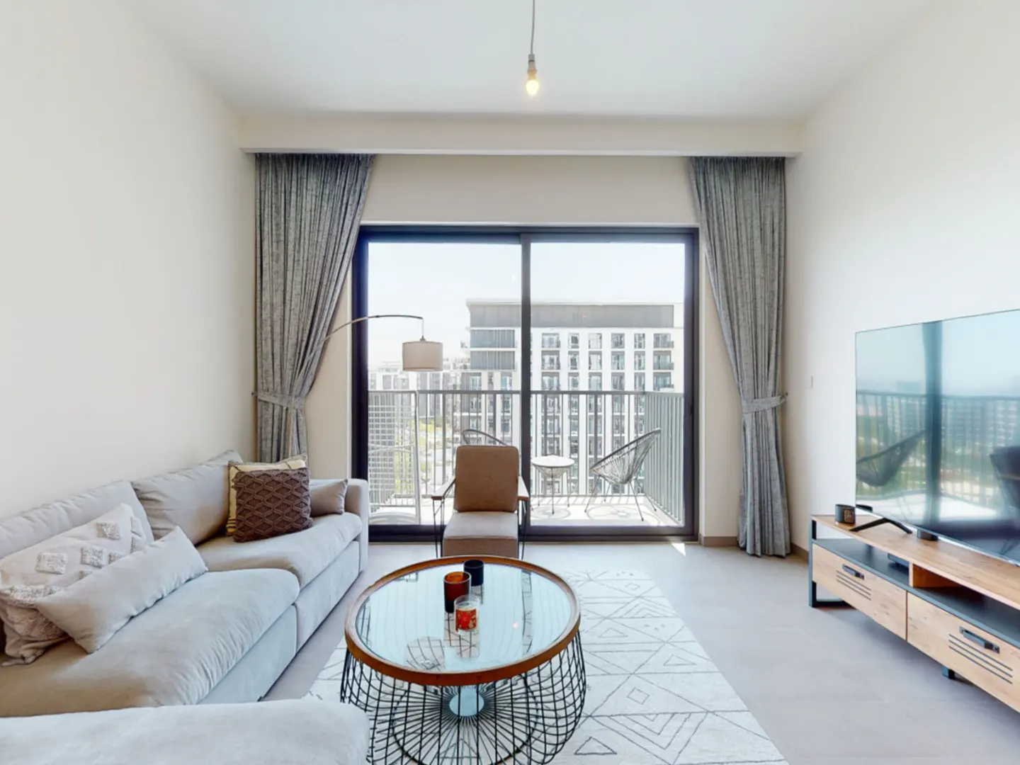 Modern living room with a grey sofa, glass coffee table, TV console, and a balcony view of city buildings.