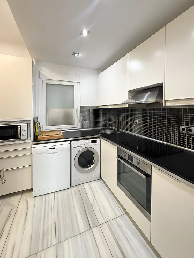 Modern kitchen with white cabinets, black tiled backsplash, marble-look floors, built-in appliances, and a washer-dryer.