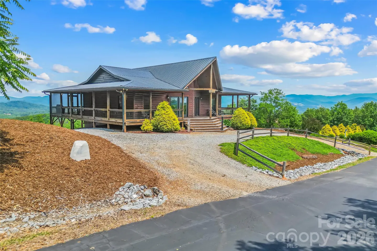 Exterior view of a brown log cabin with a metal roof, surrounded by green trees and mountains under a blue sky with white clouds.