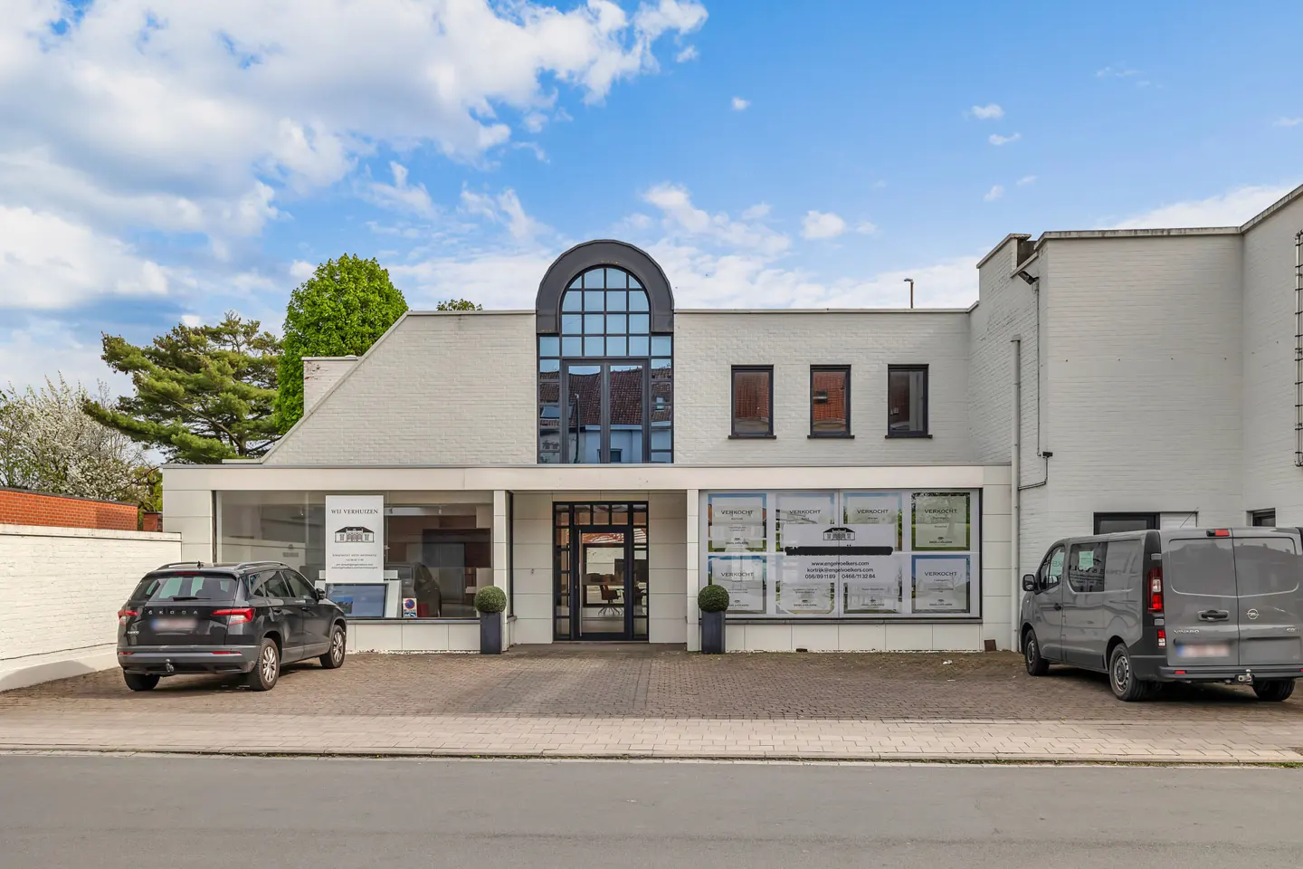 Exterior of a white brick real estate office with a black car and van parked in front. Blue sky with clouds above.