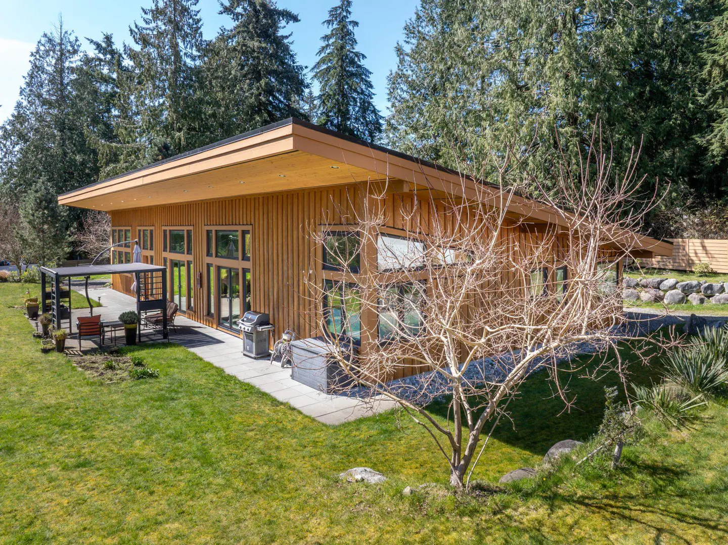 Modern wood-sided home with a flat roof, large windows, and a patio with outdoor furniture, surrounded by green lawn and trees.