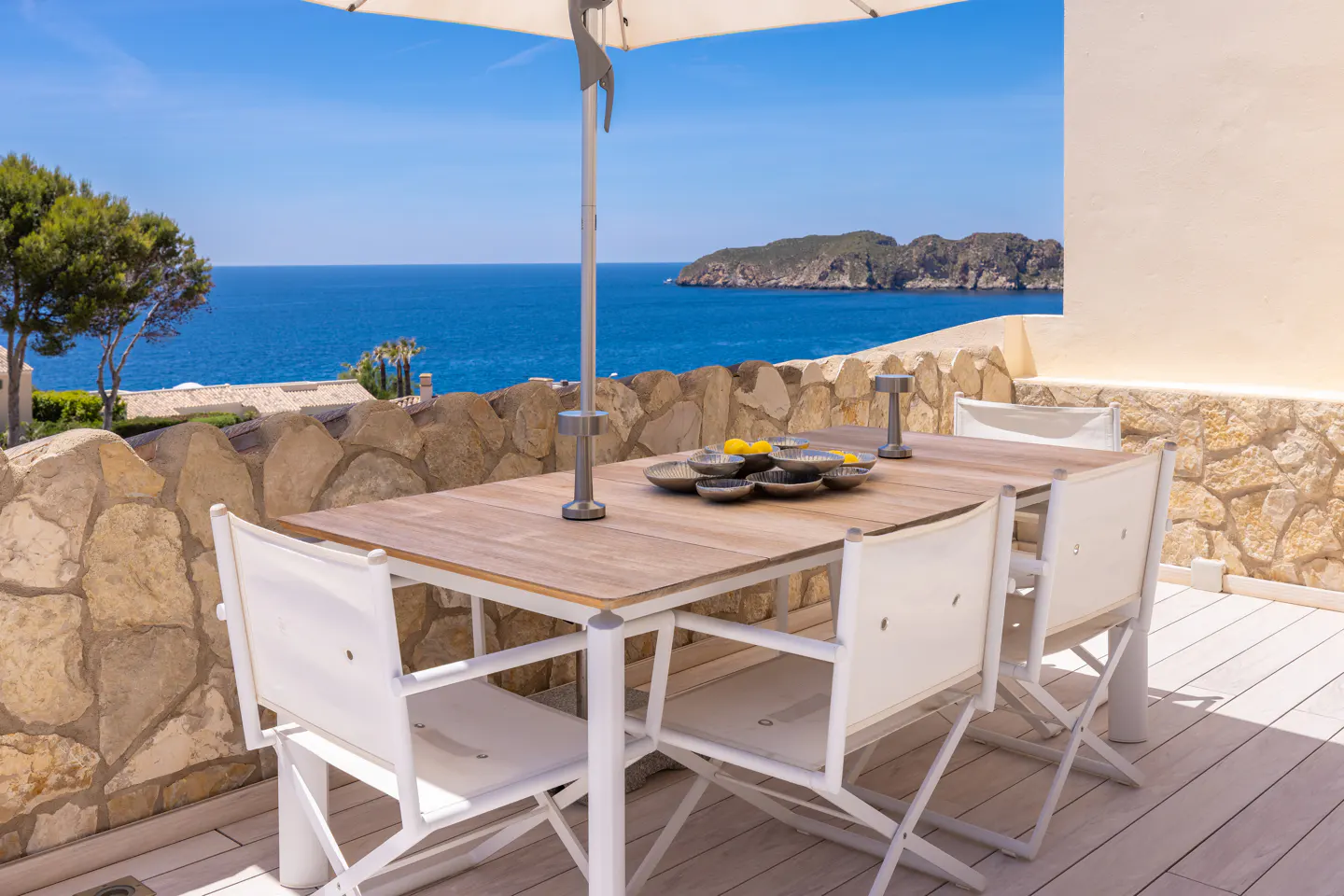Outdoor dining area with a wood table, white chairs, and umbrella overlooking the blue ocean and a rocky island.