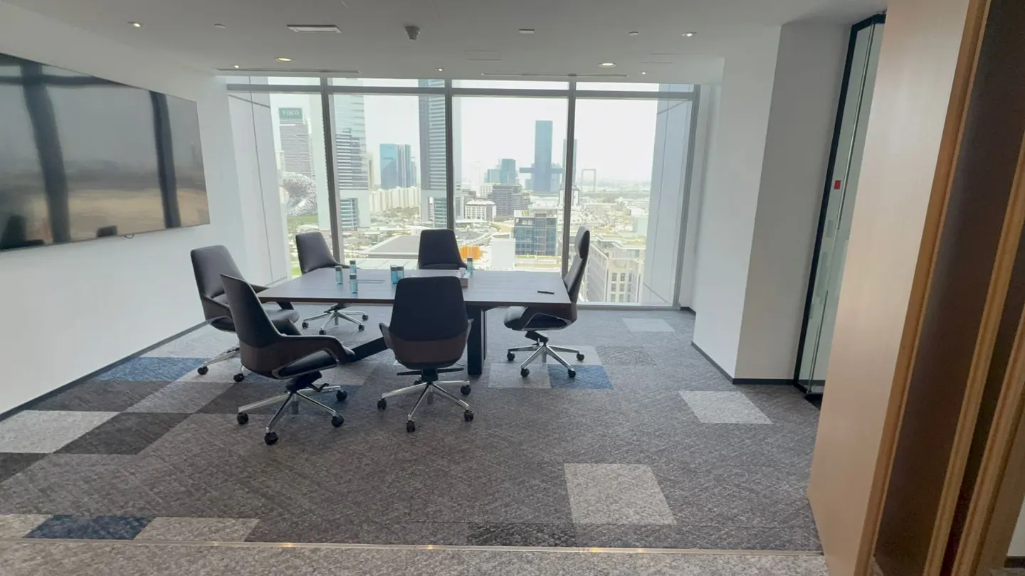 Conference room with a large window overlooking a city skyline. A dark table is surrounded by black chairs. A large screen is on the wall.