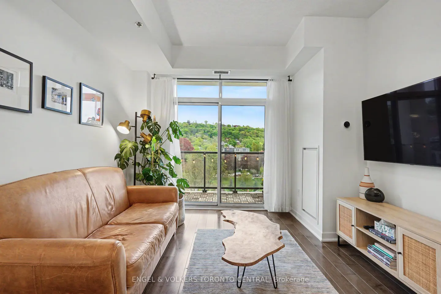 Living room with brown leather sofa, wood coffee table, TV, and balcony view of trees and water.