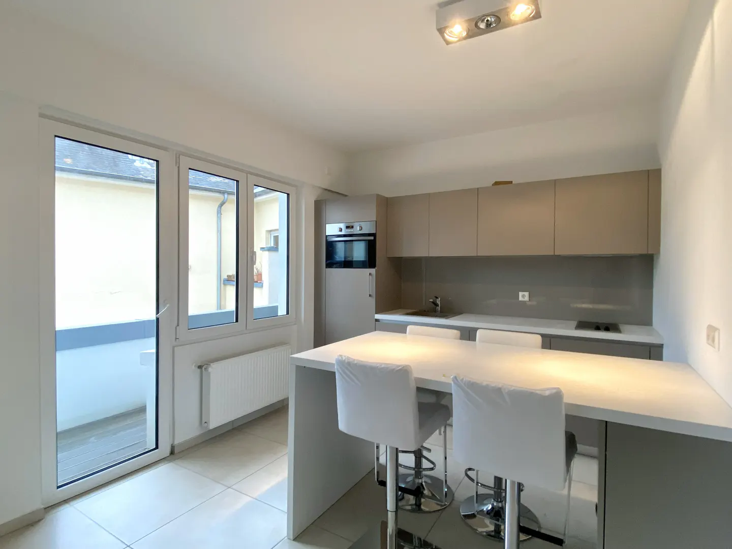 A modern kitchen with white counter, gray cabinets, and three white bar stools. A glass door leads to a balcony.