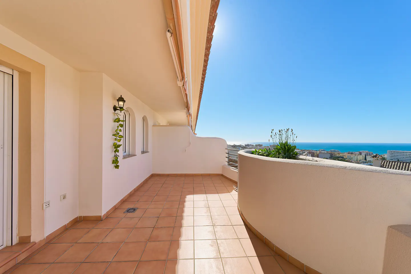 A sunlit balcony with terracotta tiles, white walls, and a curved railing overlooking a cityscape and the blue ocean.