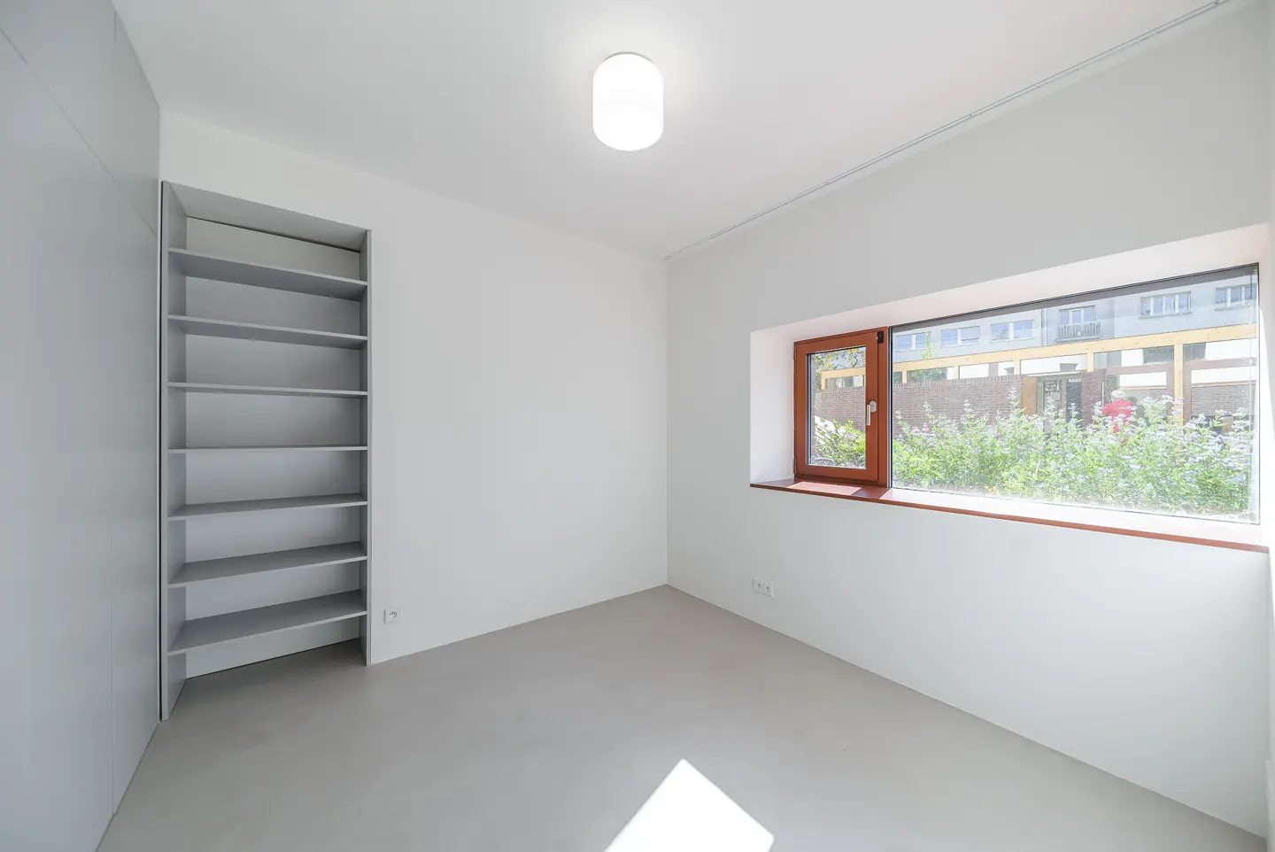 Bright, empty room with white walls, a gray floor, and a built-in gray bookshelf. A long, narrow window shows greenery outside.