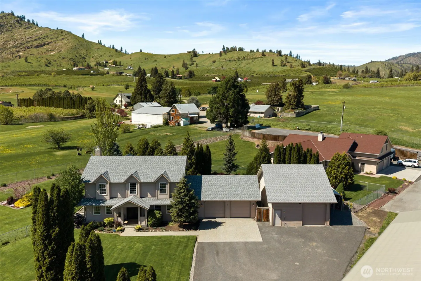 Aerial view of a gray two-story house with a three-car garage, green lawn, and a backdrop of rolling green hills.