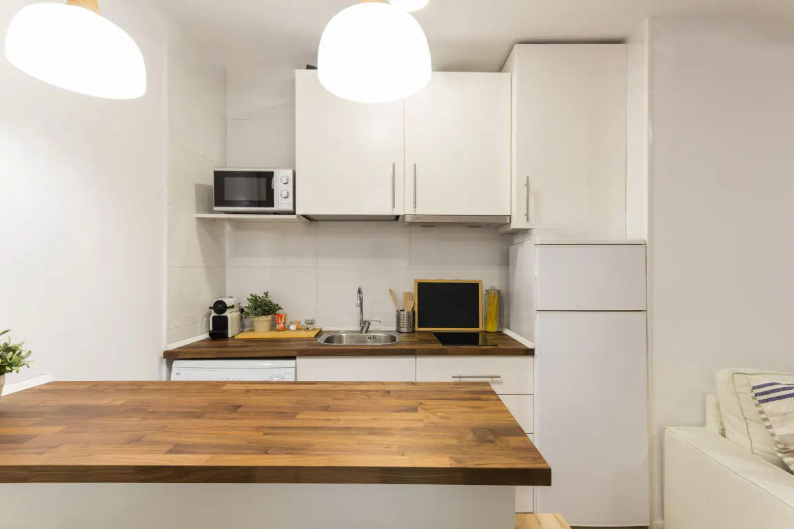 A bright, modern kitchen with white cabinets, a wood countertop, and stainless steel appliances. Overhead lighting illuminates the space.